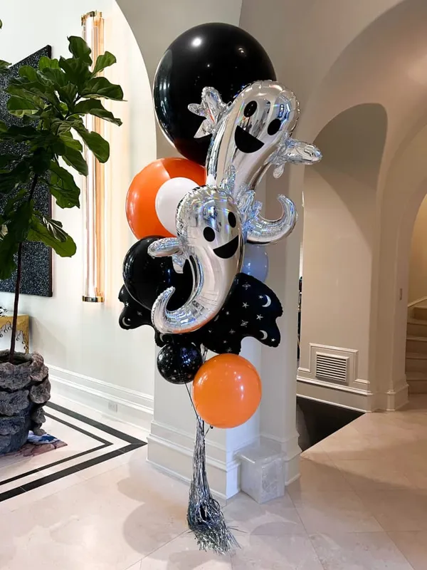 The image shows a large, whimsical balloon sculpture of a ghost-like figure surrounded by black, orange, and white balloons in a decorative indoor setting with a potted plant in the background.