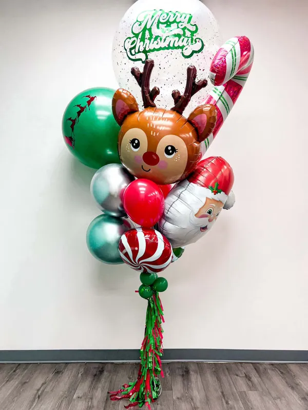 The image shows a colorful bouquet of Christmas-themed balloons, including a reindeer-shaped balloon, candy cane-shaped balloons, and a "Merry Christmas" balloon, all set against a plain white wall and wooden floor.