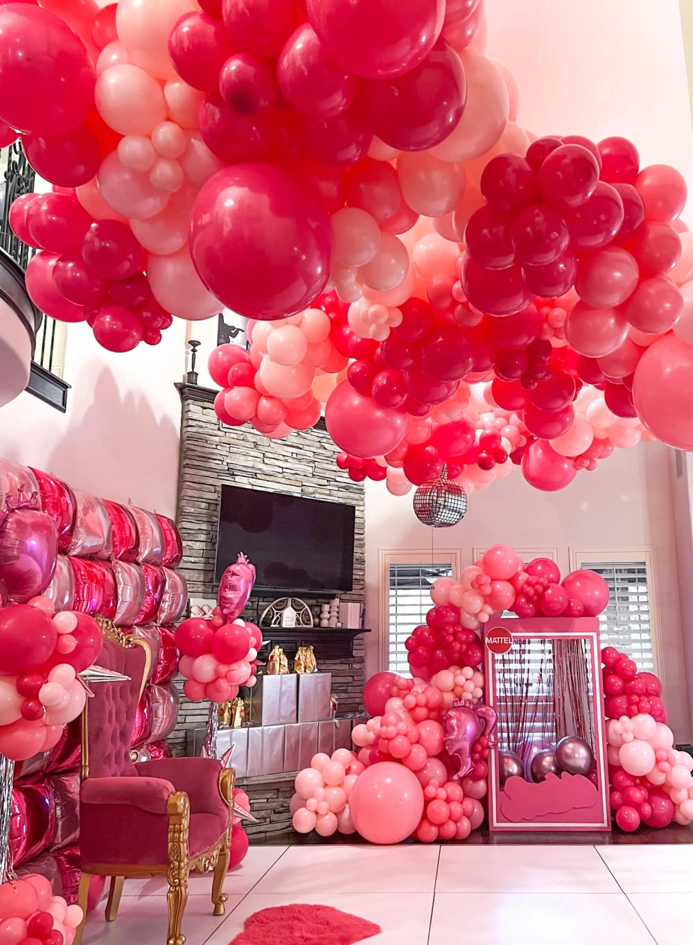 A vibrant and whimsical display of red and pink balloons fills the room, creating a festive and visually striking atmosphere. The balloons are clustered in various sizes and shapes, adorning the space and drawing the eye to the ornate furniture and decor in the background.