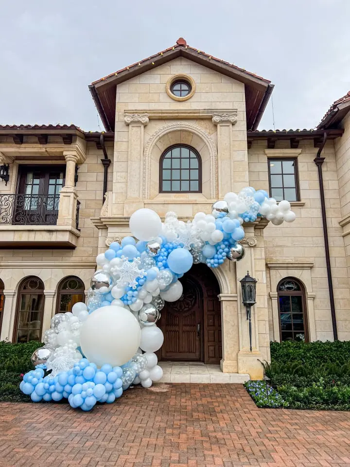 a large balloon arch with blue and white balloons in front of a large house on a brick driveway in front of a house