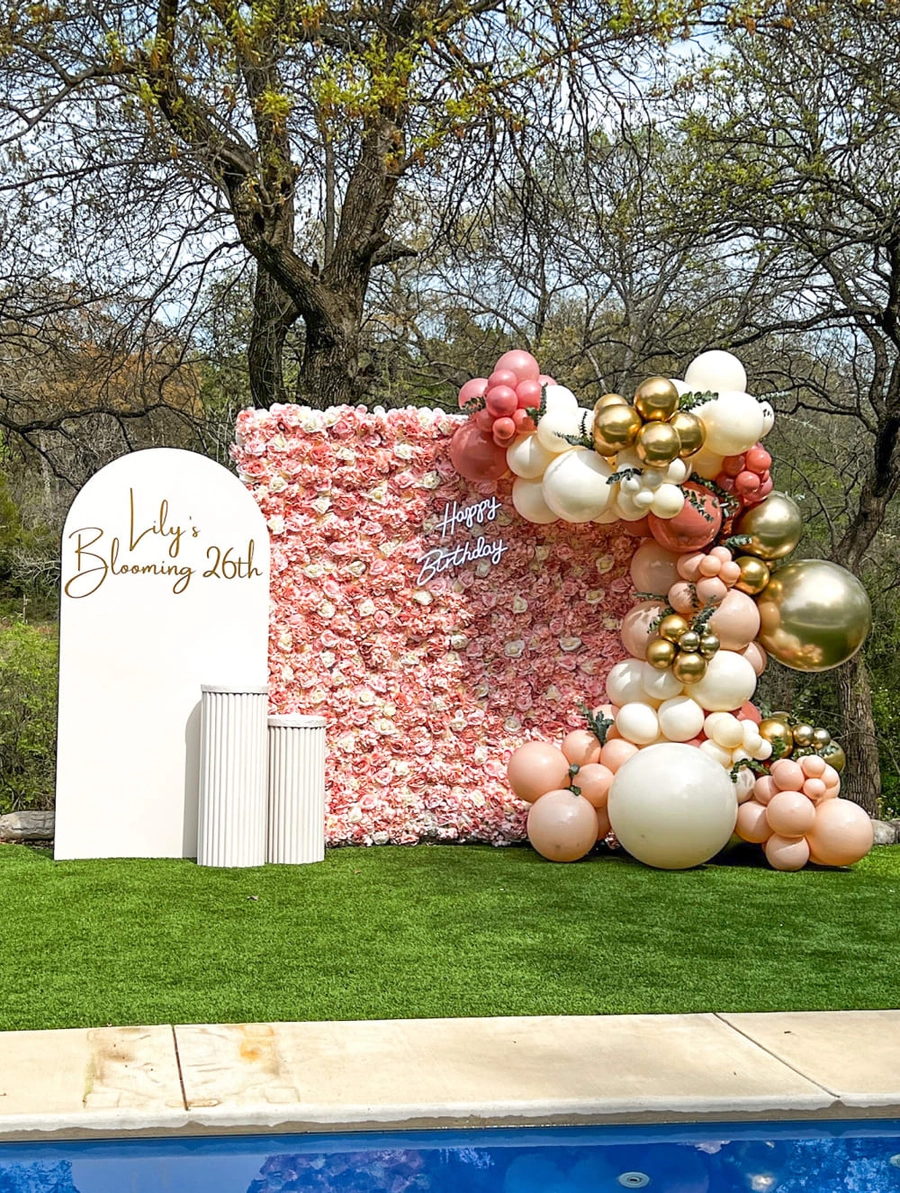 A vibrant and whimsical balloon display adorns a grassy outdoor setting, with a large floral backdrop and a sign celebrating a 26th birthday.