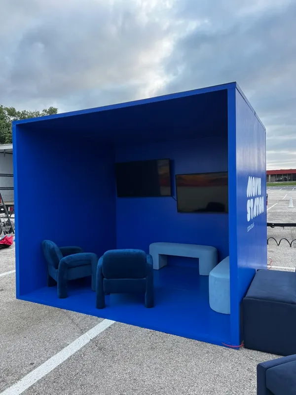 A blue and white building with a blue door is located in a parking lot, with a blue couch and a blue chair placed in front of it.