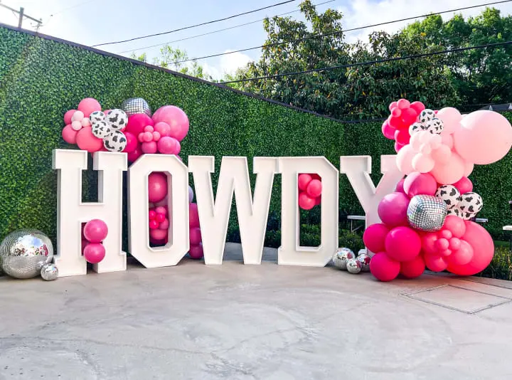 a sign that says hollywood surrounded by balloons and streamers in front of a wall of greenery with a bunch of silver and pink balloons