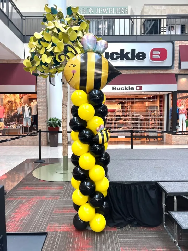 a bunch of balloons in the shape of a bee on a table in a mall with people walking by