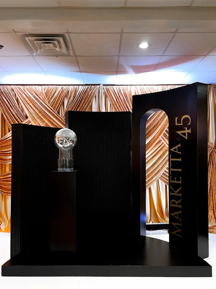 The image depicts a stage-like setup with a large black backdrop and a glass display case in the foreground, surrounded by a patterned fabric wall in warm tones.