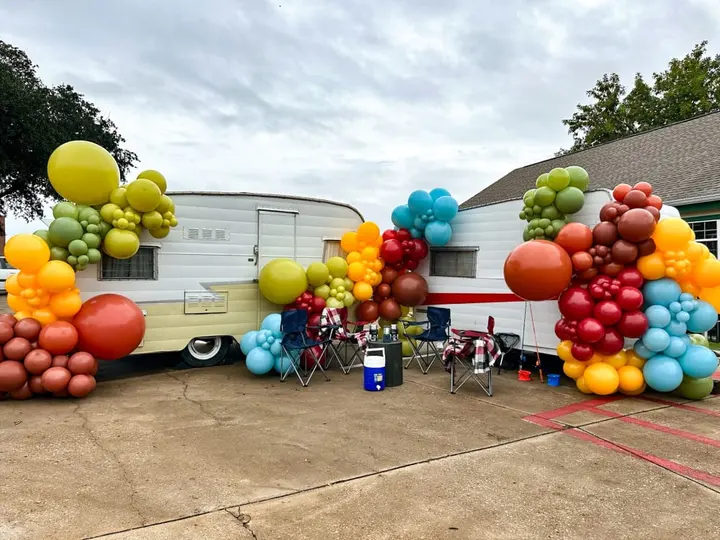 A colorful display of balloons in various shapes and sizes, including round, oblong, and elongated forms, surrounds a vintage-style camper trailer in the background, creating a vibrant and festive atmosphere.