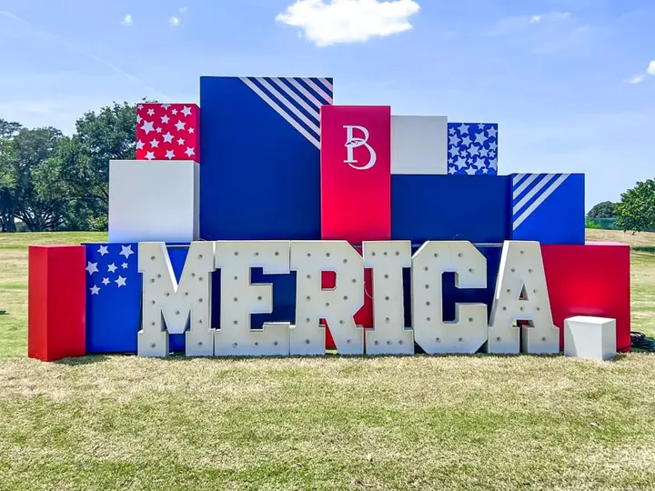 A large, colorful display featuring the word "MERICA" made up of various geometric shapes and patterns in red, white, and blue, set against a grassy field with trees in the background.