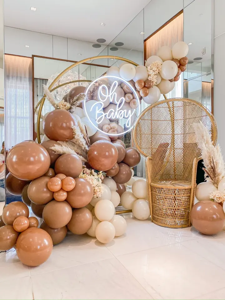 A lavish baby shower display featuring a neon sign that says "Oh Baby", surrounded by an abundance of balloons in various shades of brown and beige, along with a wicker chair and other decorative elements.