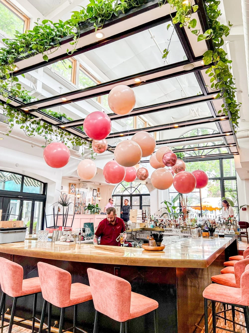 A cozy indoor dining area with a glass ceiling, featuring a wooden bar counter surrounded by pink upholstered stools and an array of colorful paper lanterns hanging overhead, with lush greenery cascading from the ceiling.