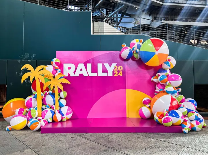 A vibrant and colorful display featuring a large "RALLY 2024" sign surrounded by inflatable beach balls, palm trees, and other playful elements against a backdrop of industrial structures.