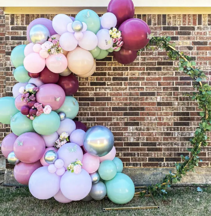 a number made out of balloons on a lawn next to a brick wall with a wreath of flowers and vines