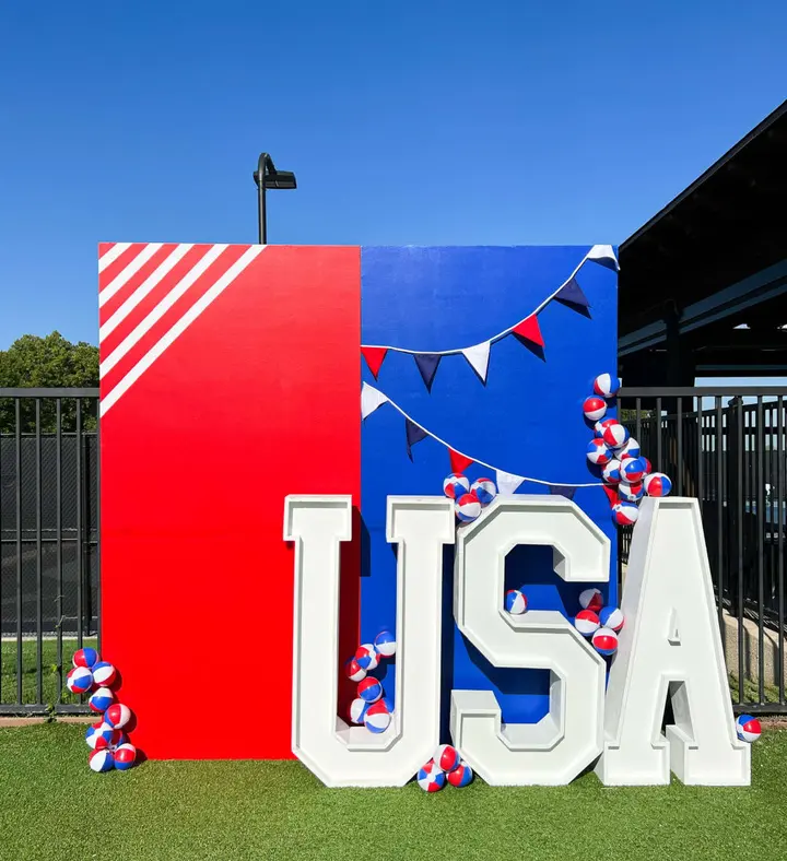 A large, colorful display featuring the letters "USA" surrounded by red, white, and blue balloons, set against a clear blue sky and a black metal fence in the background.