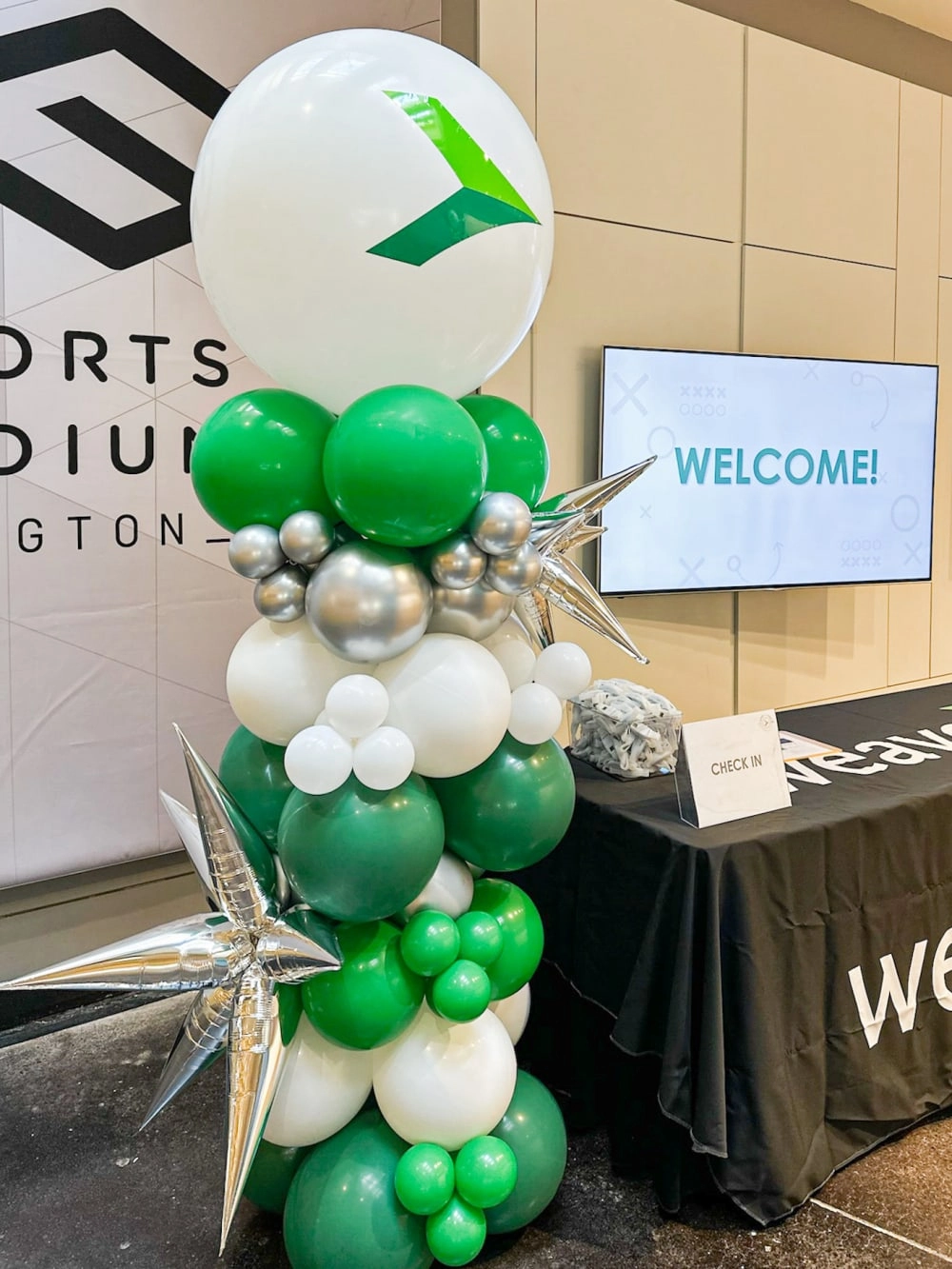 The image shows a decorative balloon arch in shades of green and white, with a large circular logo in the center, set against a backdrop of a "Welcome" sign and other event-related decor.