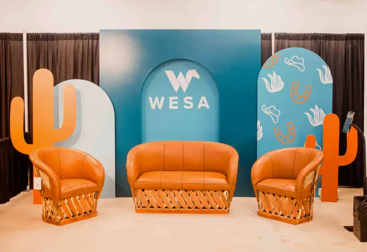 The image features a vibrant and modern setup with orange wicker chairs and a large blue backdrop displaying the "WESA" logo, surrounded by abstract shapes and patterns.