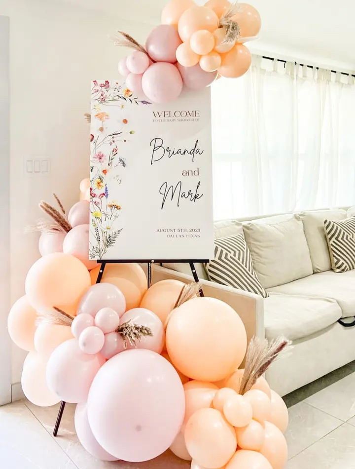 The image depicts a welcoming scene with a large floral arrangement of balloons and flowers in soft, pastel colors, set against a cozy living room backdrop with a sofa and curtains.