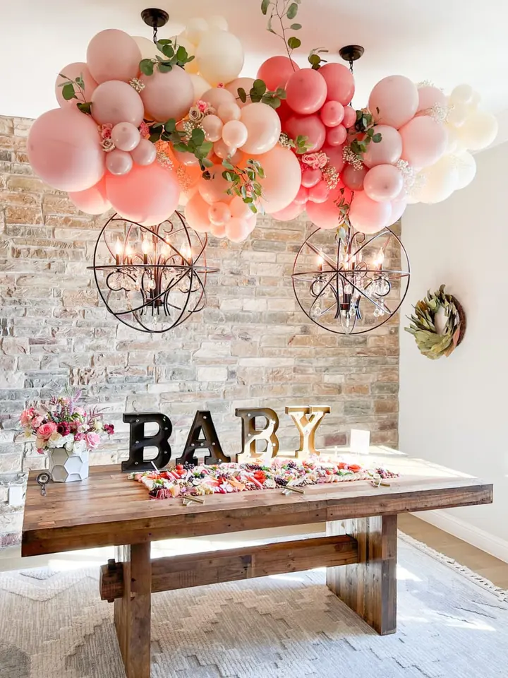 A rustic wooden table adorned with a variety of pink and white balloons, surrounded by a brick wall and complemented by two industrial-style light fixtures, creating a warm and festive atmosphere.