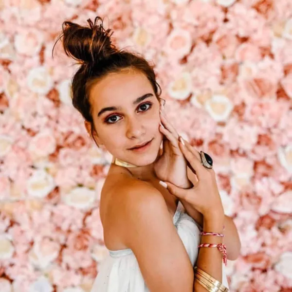 a woman posing for a picture in front of a wall of flowers with her hand on her cheek and her hand under her chin
