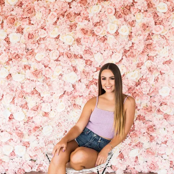 a woman sitting on a chair in front of a wall of pink flowers with a smile on her face