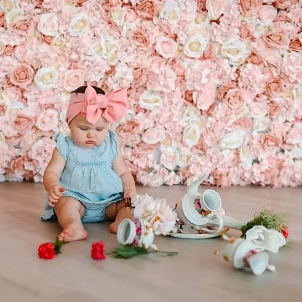 a baby girl sitting on the floor next to a wall of flowers and a tea pot with a pink bow on it
