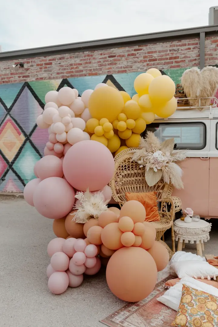 A vibrant display of colorful balloons and decorative elements in the foreground, set against a backdrop of a brick wall with a mural.