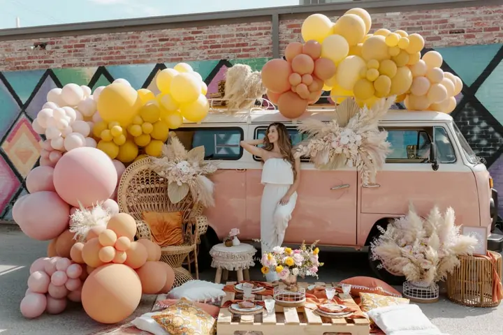 A colorful and whimsical scene featuring a vintage pink van adorned with an abundance of yellow, pink, and white balloons, surrounded by various decorative elements and a festive display.