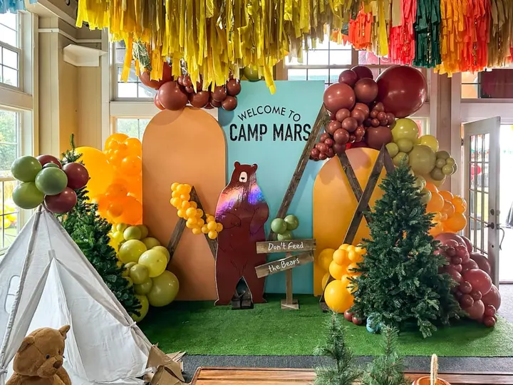 A colorful and whimsical camp-themed display with various decorative elements, including hanging streamers, balloons, and a large welcome sign featuring a bear character, set against a backdrop of lush greenery and a window.