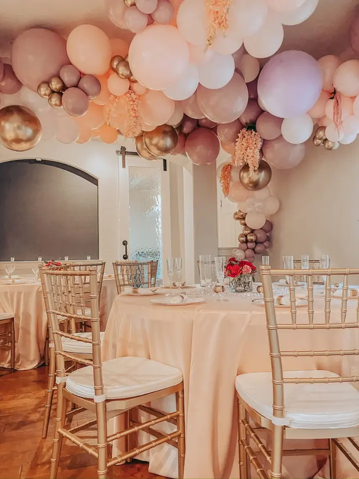 A festive event space with a table set for a celebration, adorned with a vibrant balloon arch in shades of pink, peach, and gold, creating a warm and inviting atmosphere.