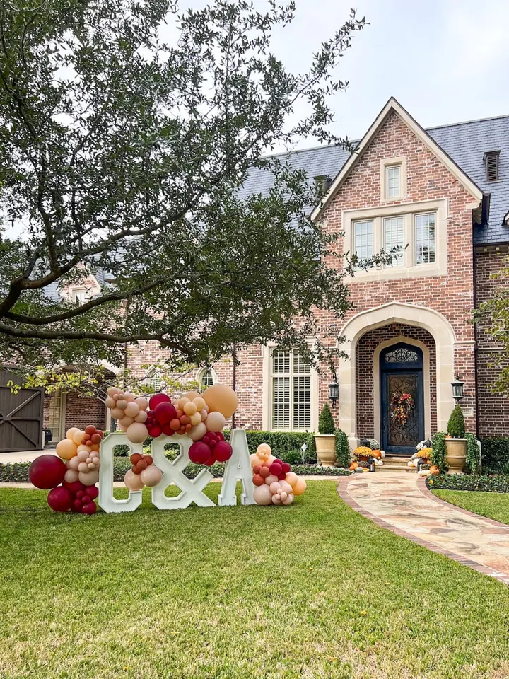 A large, brick house with a decorative entryway is surrounded by a well-manicured lawn, featuring a colorful display of oversized balloons and the word "Bday" in the foreground.