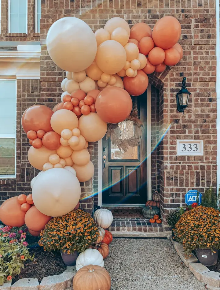 A vibrant display of orange and white balloons adorns the entrance of a brick building, complemented by potted plants and pumpkins on the porch, creating a festive autumn atmosphere.