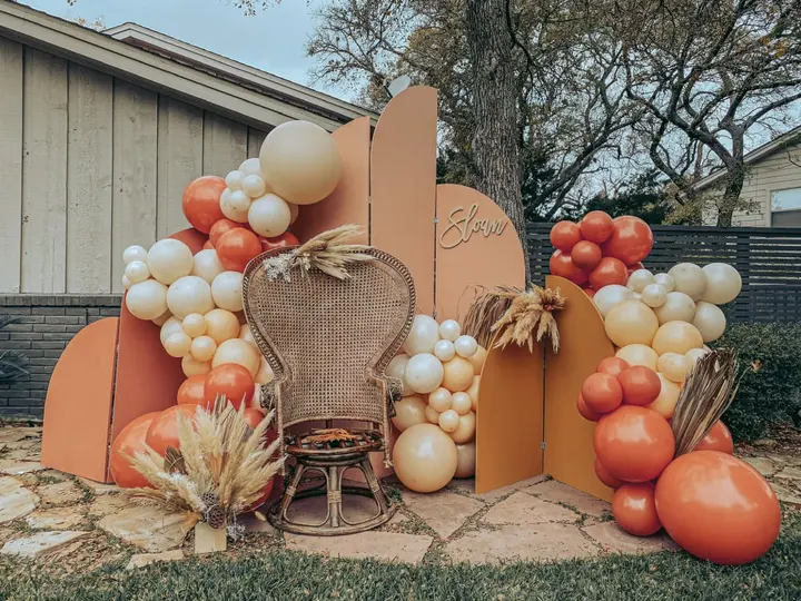 A rustic, autumn-themed display featuring an assortment of decorative elements, including balloons, pumpkins, and woven baskets, set against a backdrop of a wooden structure and lush foliage.