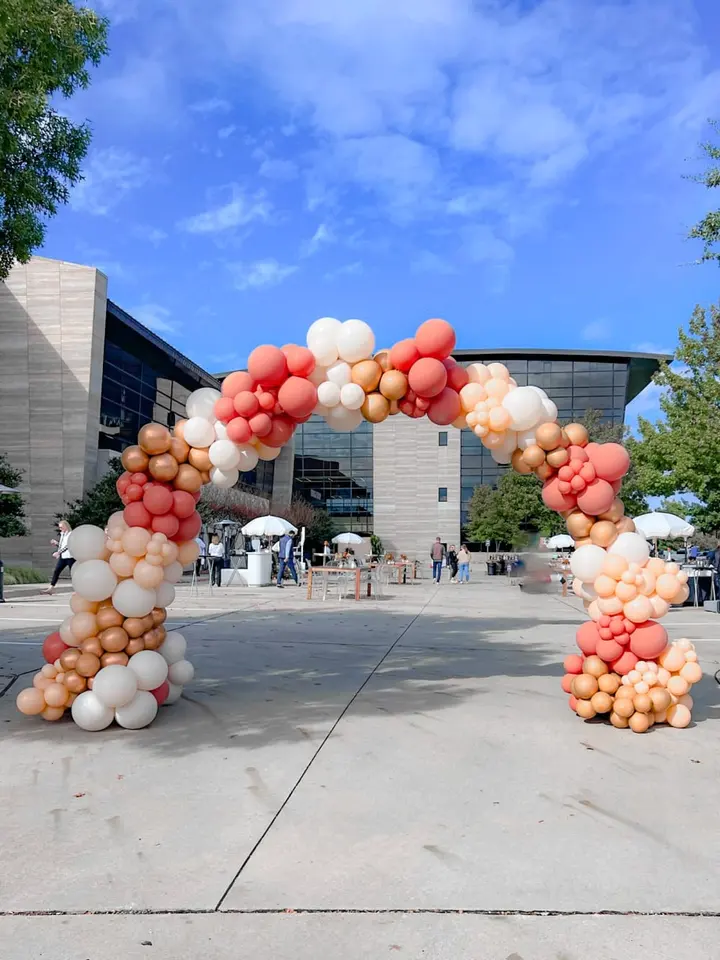 A large, colorful balloon arch stands in the foreground, framing the entrance to a modern, glass-walled building with a blue sky and scattered clouds in the background.