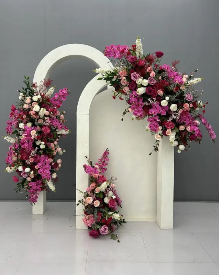 A white arched structure adorned with an abundance of vibrant pink and purple flowers, set against a plain gray background, creating a visually striking and elegant floral display.