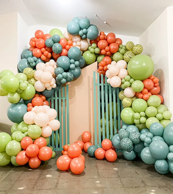 A vibrant and colorful balloon arch made up of various shades of green, orange, and blue balloons, creating a visually striking and festive display in a minimalist indoor setting.