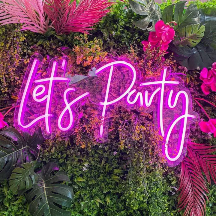 a pink neon sign that says let's party surrounded by plants and flowers on a wall of greenery