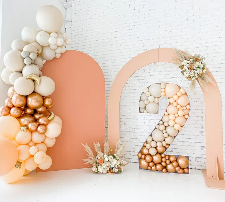 The image features a decorative arrangement of balloons and floral elements in shades of peach, orange, and white, creating a visually appealing and festive display against a plain white brick wall backdrop.