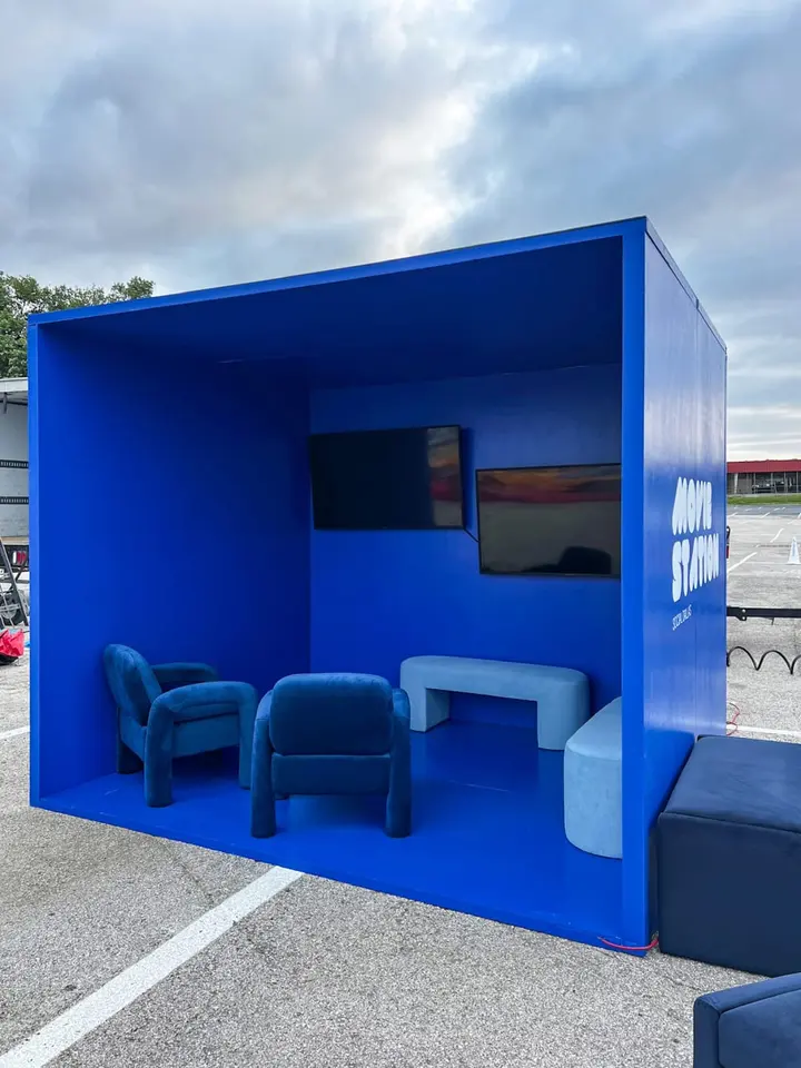 A bright blue modular structure with seating and a television screen, situated in an outdoor setting with a cloudy sky in the background.