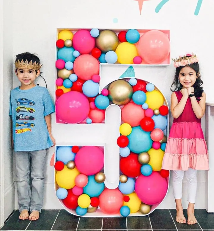 a group of children standing next to a large number 50 balloon wall in a room with a white wall and black tile floor