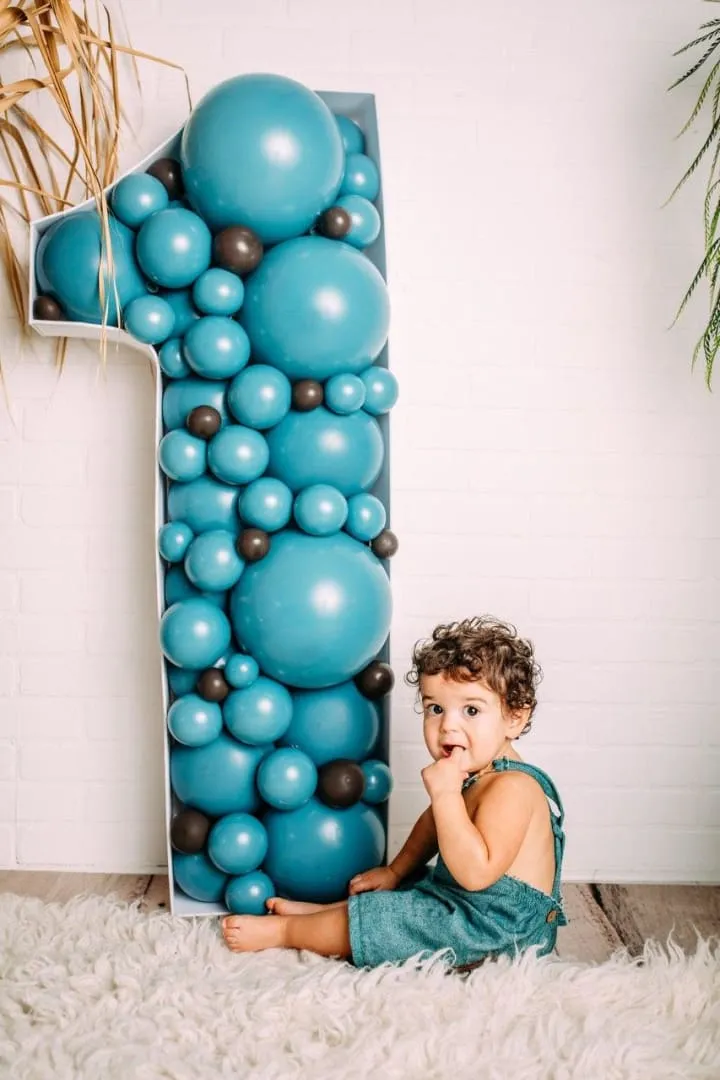 a little boy sitting on the floor next to a number one made out of balloons and a balloon garland