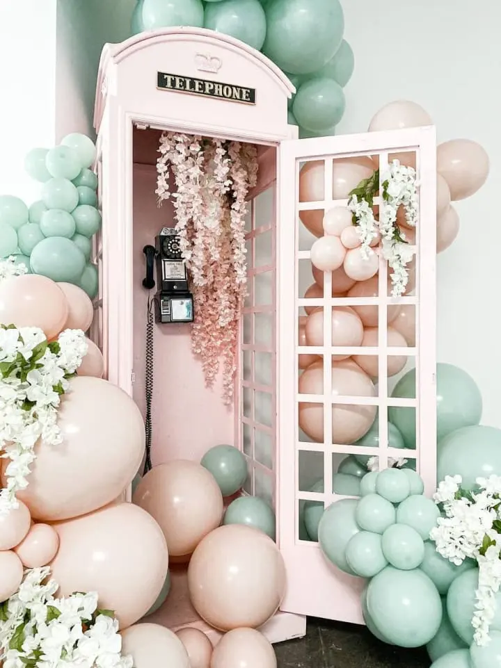 a pink phone booth surrounded by balloons, flowers, and a bunch of balloons in a room with a phone on the wall