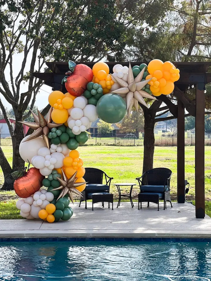 A vibrant and whimsical balloon arch adorns the outdoor space, featuring a variety of colorful shapes and sizes, creating a festive and inviting atmosphere. In the background, a lush green lawn and a swimming pool can be seen, adding to the serene and relaxing ambiance of the scene.