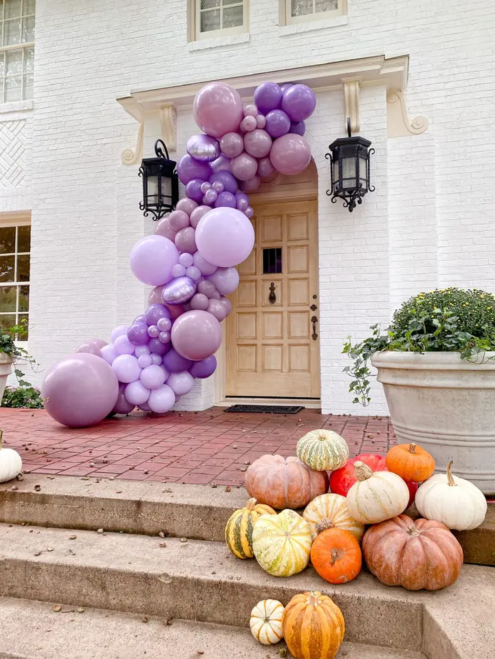 The image shows a grand entrance to a building, adorned with a vibrant display of purple and pink balloons cascading down the steps, complemented by an assortment of colorful pumpkins and gourds arranged in the foreground.