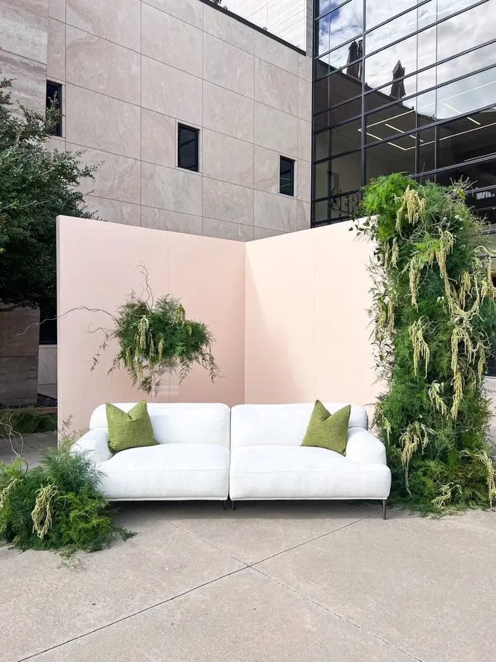 A modern, minimalist outdoor seating area with a white sofa surrounded by lush greenery and a concrete building in the background.