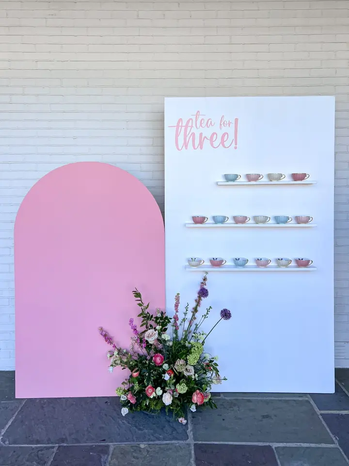 A large pink backdrop stands in the foreground, with a colorful floral arrangement placed in front of it. In the background, a white wall displays a sign that reads "Tea for three!" and features a display of various teacups.