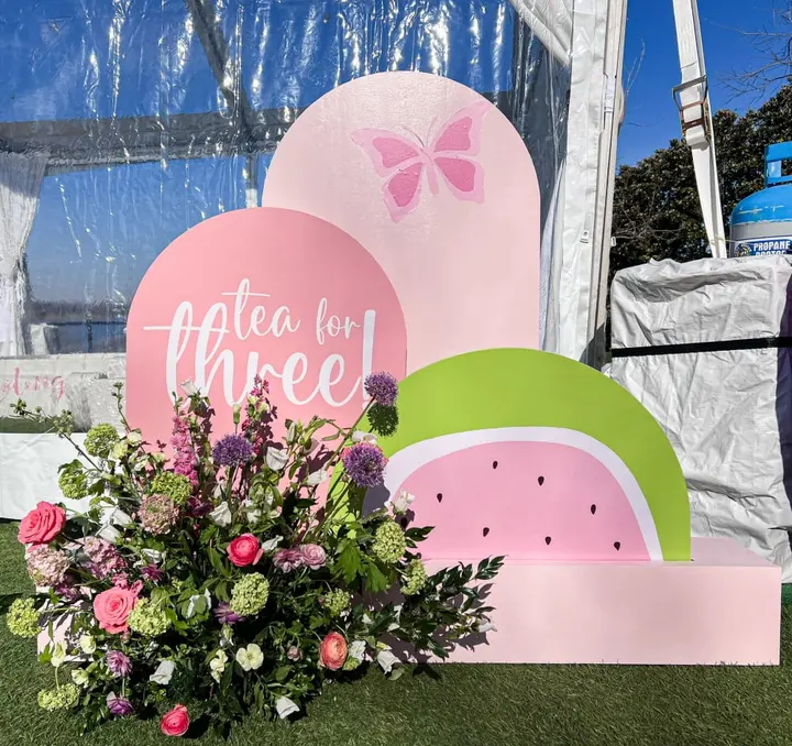 The image shows a display of large pink and green shapes resembling watermelons and butterflies, with a bouquet of colorful flowers in the foreground, set against a backdrop of a building and blue sky.