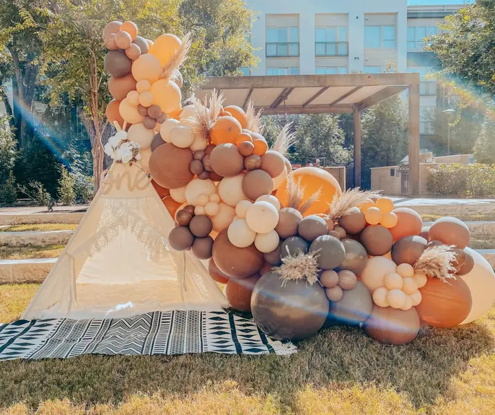 A vibrant and whimsical display of balloons in various shades of orange, brown, and white, set against a backdrop of a building and lush greenery.