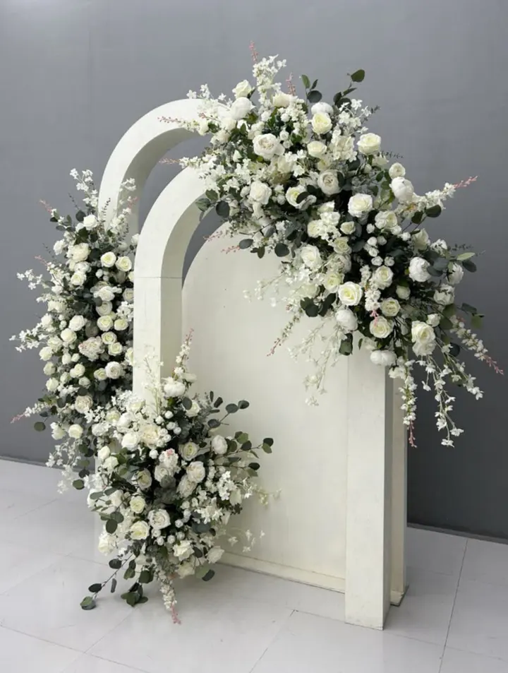 A white, arched floral arrangement filled with delicate white flowers and greenery sits on a plain white surface against a gray background.