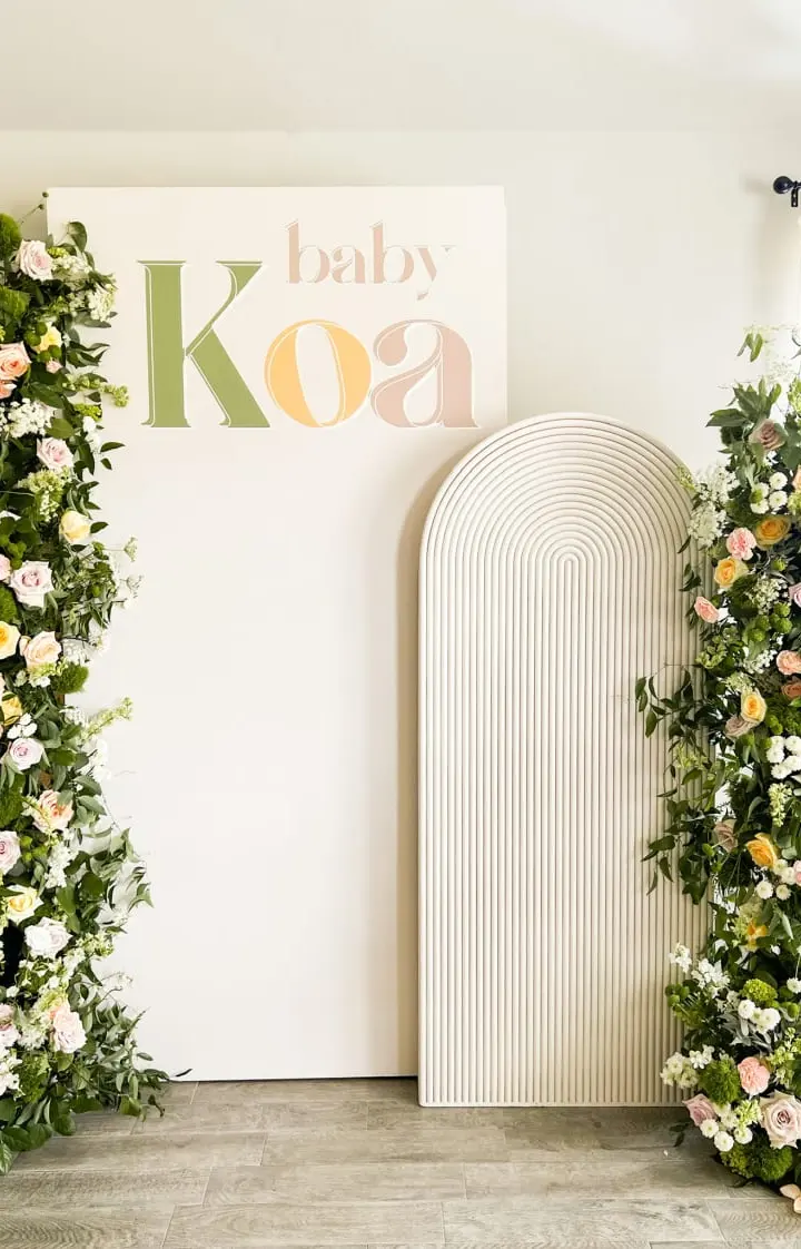 A floral archway adorned with lush greenery and vibrant flowers frames a sign that reads "Baby Koa", set against a neutral-toned background.