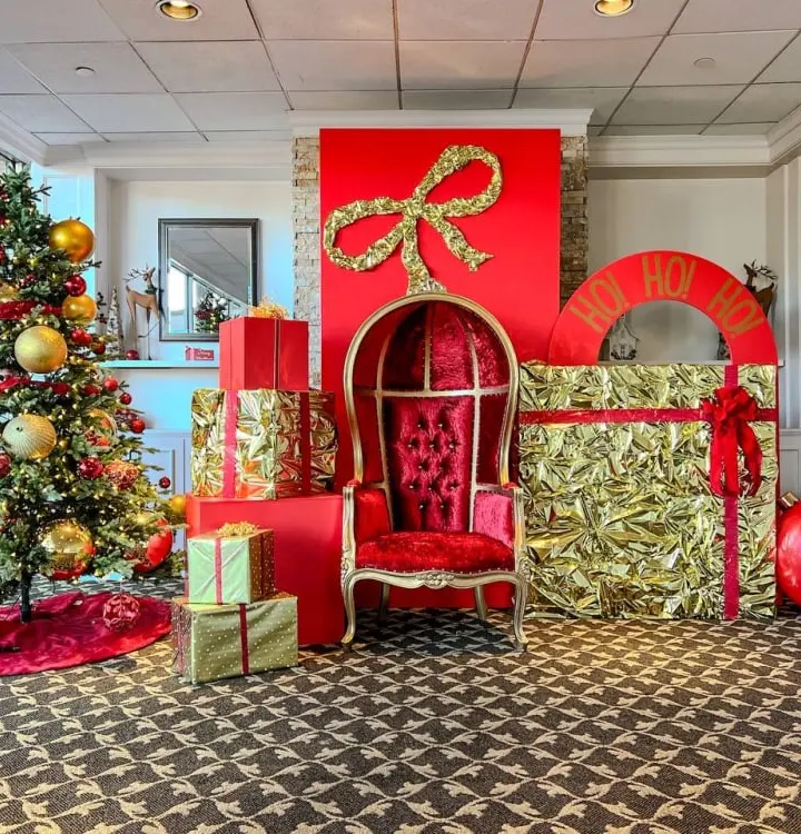 A festively decorated room with a Christmas tree, ornaments, and a large red and gold throne-like chair surrounded by wrapped gifts and other holiday decor.
