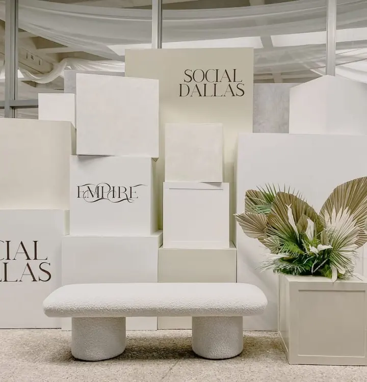 The image depicts a minimalist and modern display setup, featuring white podiums and pedestals with the text "Social Dallas" prominently displayed. The background is adorned with draped fabric, creating a visually appealing and cohesive aesthetic.