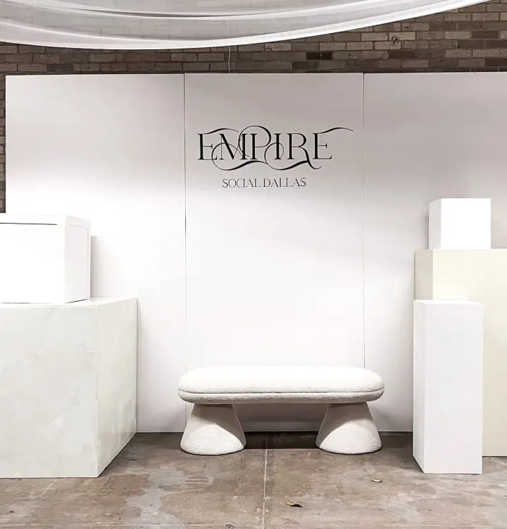 A minimalist display setup with a large white backdrop featuring the "Empire" logo, surrounded by various white pedestals and platforms on a tiled floor.
